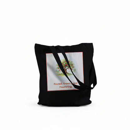 A black canvas tote bag with black shoulder straps, featuring a square graphic print on the front. The print shows a stylized tree with green and orange leaves 