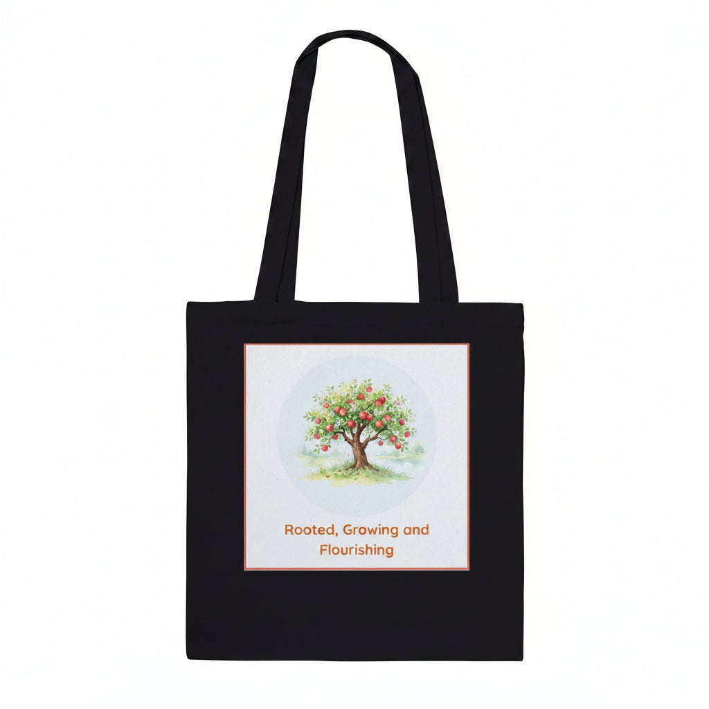A black canvas tote bag with two shoulder straps, featuring a square graphic print on the front. The print shows a watercolor-style apple tree with red apples under a light blue circle, with the text 'Rooted, Growing and Flourishing' below in orange.