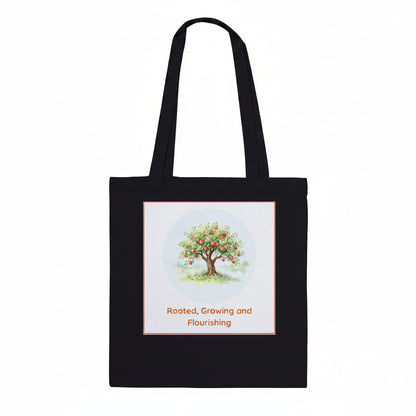 A black canvas tote bag with two shoulder straps, featuring a square graphic print on the front. The print shows a watercolor-style apple tree with red apples under a light blue circle, with the text 'Rooted, Growing and Flourishing' below in orange.