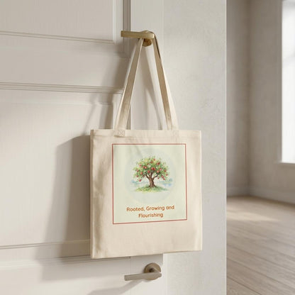 A cream canvas tote bag with a watercolor tree illustration and the text 'Rooted, Growing and Flourishing' hanging from a brass doorknob on a white paneled door.