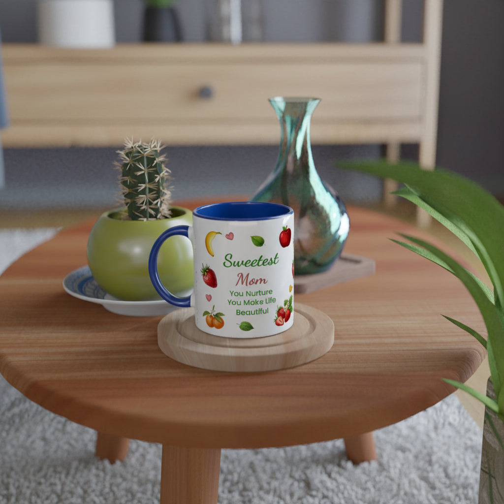 A white ceramic mug with a blue handle and rim sits on a round wooden coaster on a light wood table. The mug is printed with the words 'Sweetest Mom' and a quote, decorated with colorful illustrations of strawberries, bananas, and cherries.