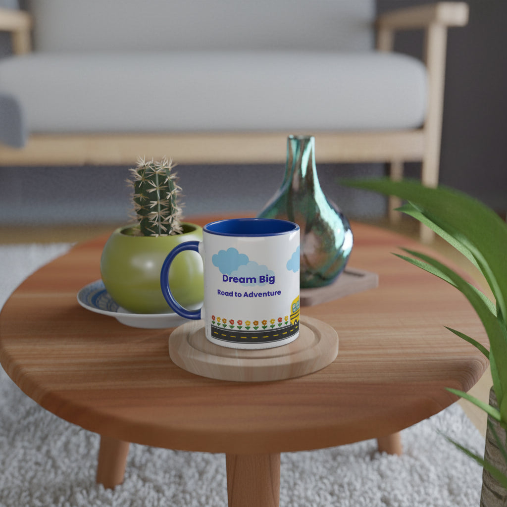 A white ceramic mug with a blue handle and interior sits on a wooden coaster on a round wooden table. The mug is printed with the words 'Dream Big Road to Adventure' in blue, along with small blue clouds and a colorful road pattern near the bottom.