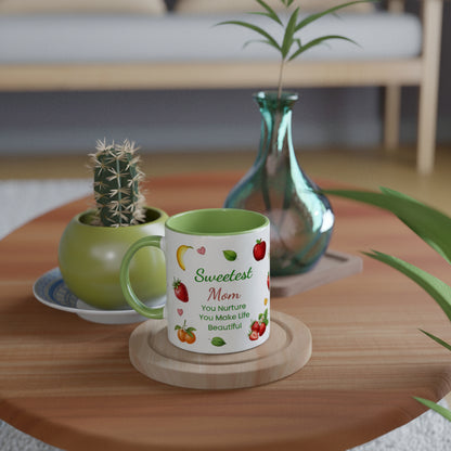 A white ceramic mug with a green handle and interior, printed with the words 'Sweetest Mom You Nurture You Make Life Beautiful' and small fruit illustrations, sits on a round wooden coaster on a wooden table. A small potted cactus and a blue-green glass vase are in the background in a softly blurred home setting.