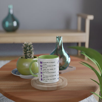 A white ceramic mug with a green handle and interior, printed with the word 'MOTHER' in purple letters and a quote, displayed on a wooden coaster on a round wooden table. The background is a softly blurred home interior with a small cactus in a green pot and decorative glass bottles.