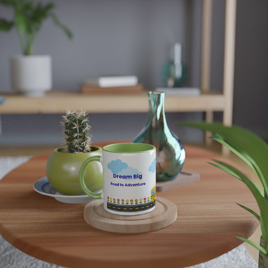 A white ceramic mug with a green handle and interior, printed with 'Dream Big Read to Adventure' and a small colorful illustration, sitting on a round wooden coaster on a wooden table in a styled room setting.