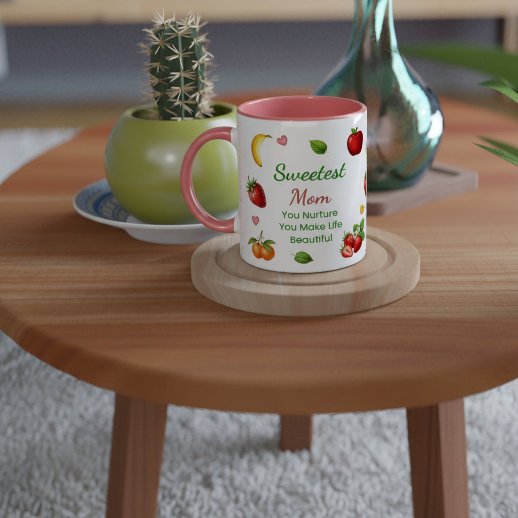 A white ceramic mug with a pink interior and handle, printed with the words 'Sweetest Mom You Nurture You Make Life Beautiful' and small fruit illustrations, sitting on a round wooden coaster on a wooden table.