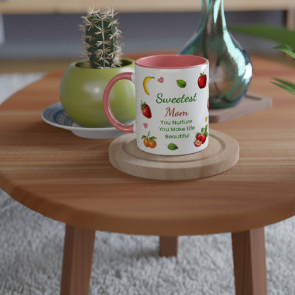 A white ceramic mug with a pink interior and handle, printed with the words 'Sweetest Mom You Nurture You Make Life Beautiful' and small fruit illustrations, sitting on a round wooden coaster on a wooden table.