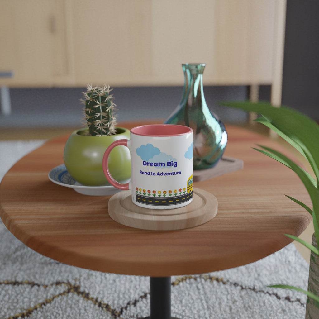 A white ceramic mug with a pink handle and interior sits on a round wooden table in a styled indoor setting. The mug is printed with blue clouds, the words 'Dream Big Road to Adventure,' and a small colorful illustration.