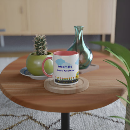 A white ceramic mug with a pink handle and interior sits on a round wooden table in a styled indoor setting. The mug is printed with blue clouds, the words 'Dream Big Road to Adventure,' and a small colorful illustration.