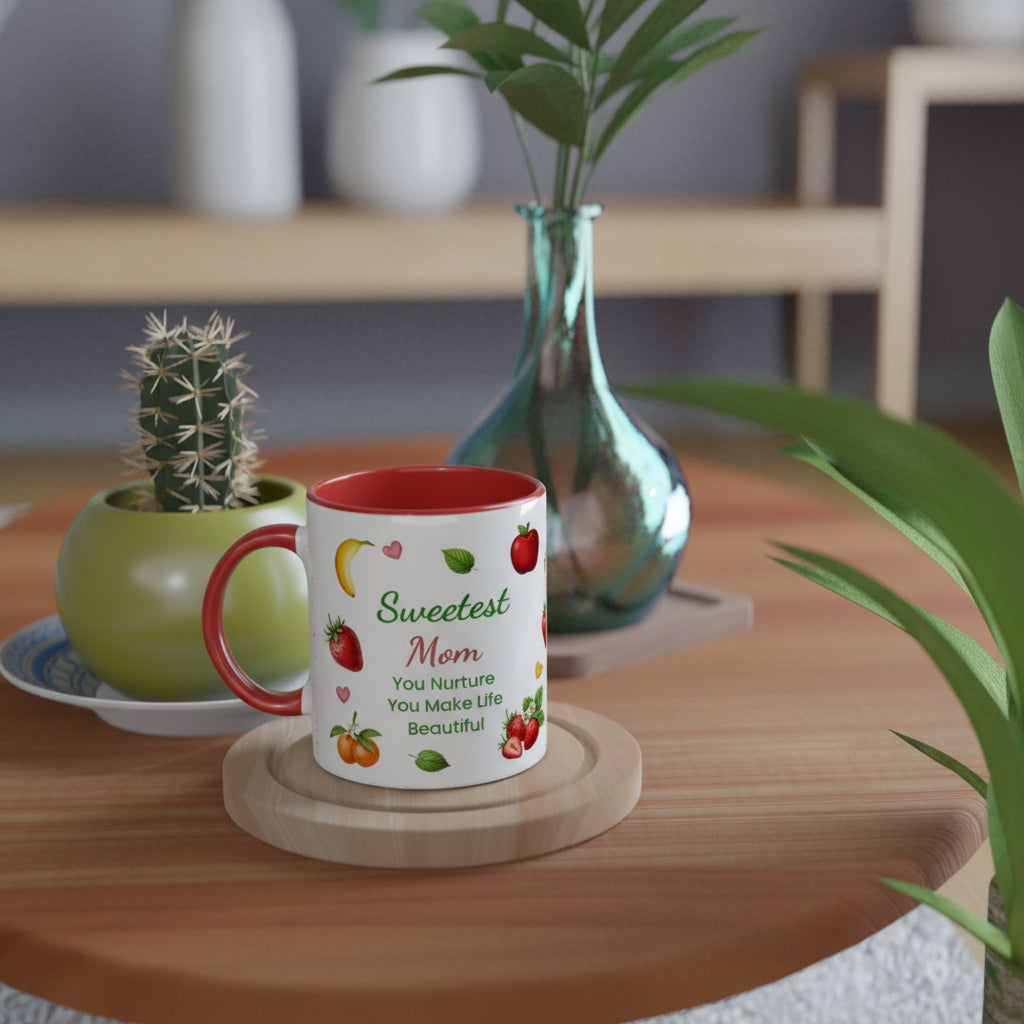 A white ceramic mug with a red interior and handle, printed with the phrase 'Sweetest Mom' and small fruit illustrations, displayed on a wooden coaster on a round wooden table in a styled home setting.