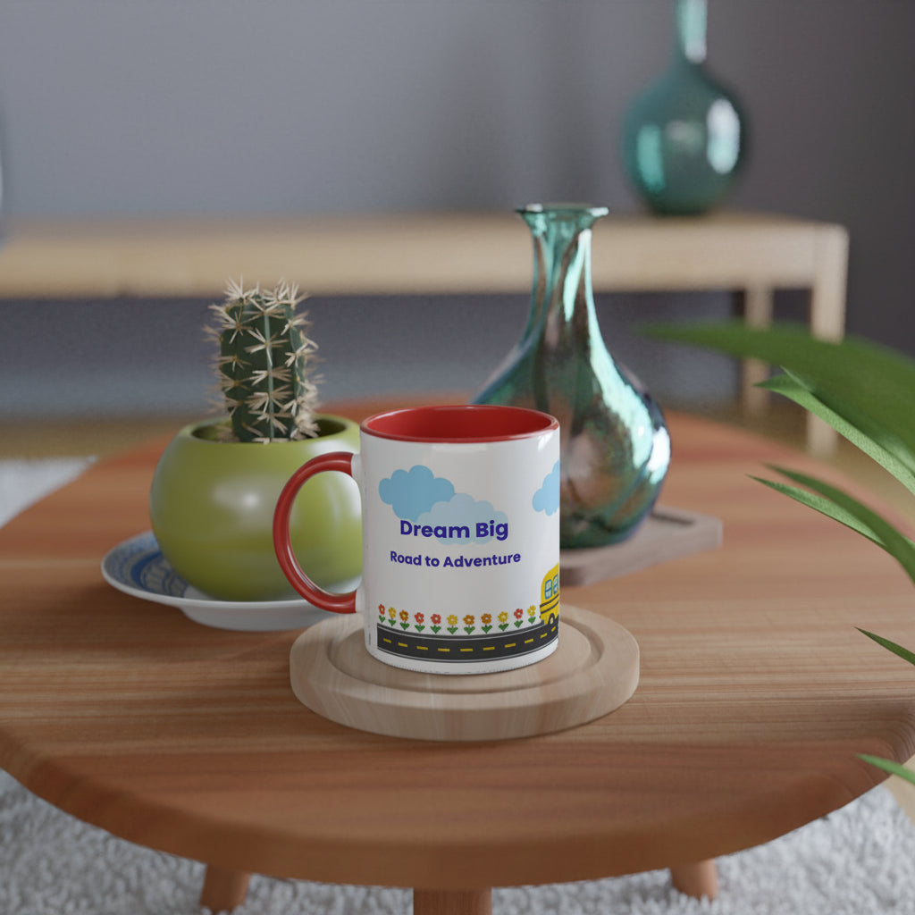 A white ceramic mug with a red handle and red interior sits on a round wooden coaster on a wooden table. The mug is printed with blue clouds, the words 'Dream Big Road to Adventure', and a small colorful illustration.