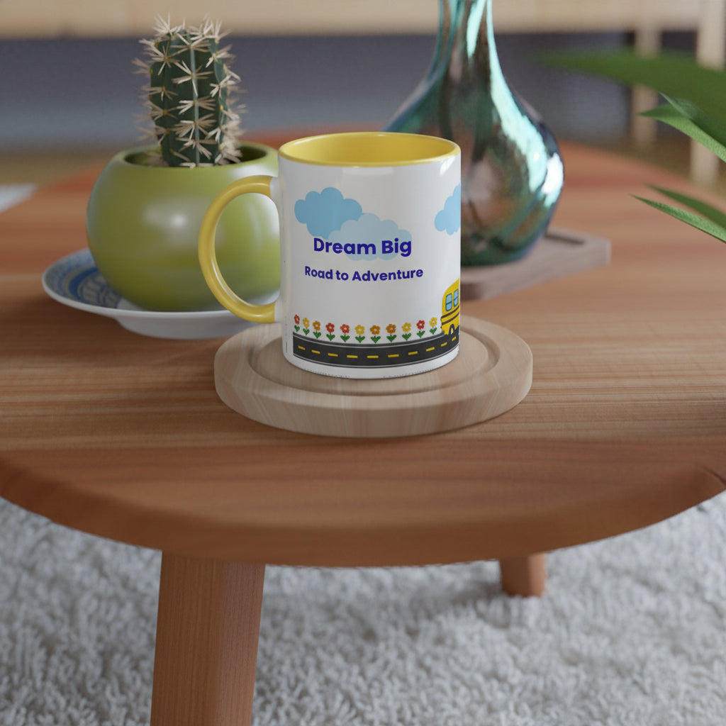 A white ceramic coffee mug with a yellow handle and interior, printed with 'Dream Big Road to Adventure' and a small bus illustration, sitting on a round wooden coaster on a wooden table.