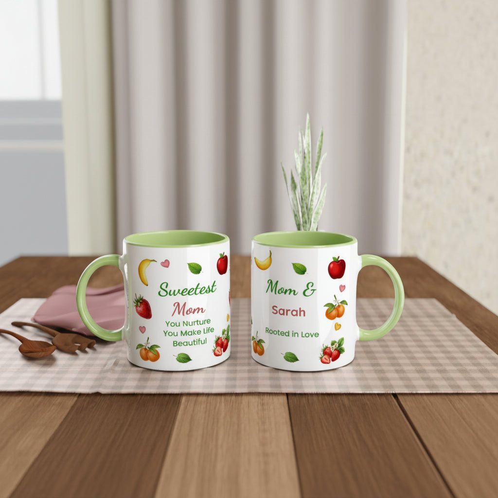 Two personalized white ceramic mugs with light green interiors and handles, displayed side by side on a wooden table. The mug on the left reads 'Sweetest Mom You Nurture You Make Life Beautiful' with illustrations of strawberries, bananas, and apples.
