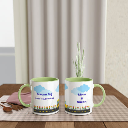 Two personalized ceramic mugs with light green interiors and handles, displayed on a wooden table. One mug reads 'Dream Big Road to Adventure' and the other reads 'Mom & Sarah', both featuring blue clouds, a yellow sun, and a multicolor road pattern at the bottom.
