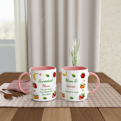 Two personalized ceramic mugs with pink interiors and handles, displayed on a wooden table. One mug is printed with 'Sweetest Mom You Nurture You Make Life Beautiful' with strawberries, bananas, and green leaves.
