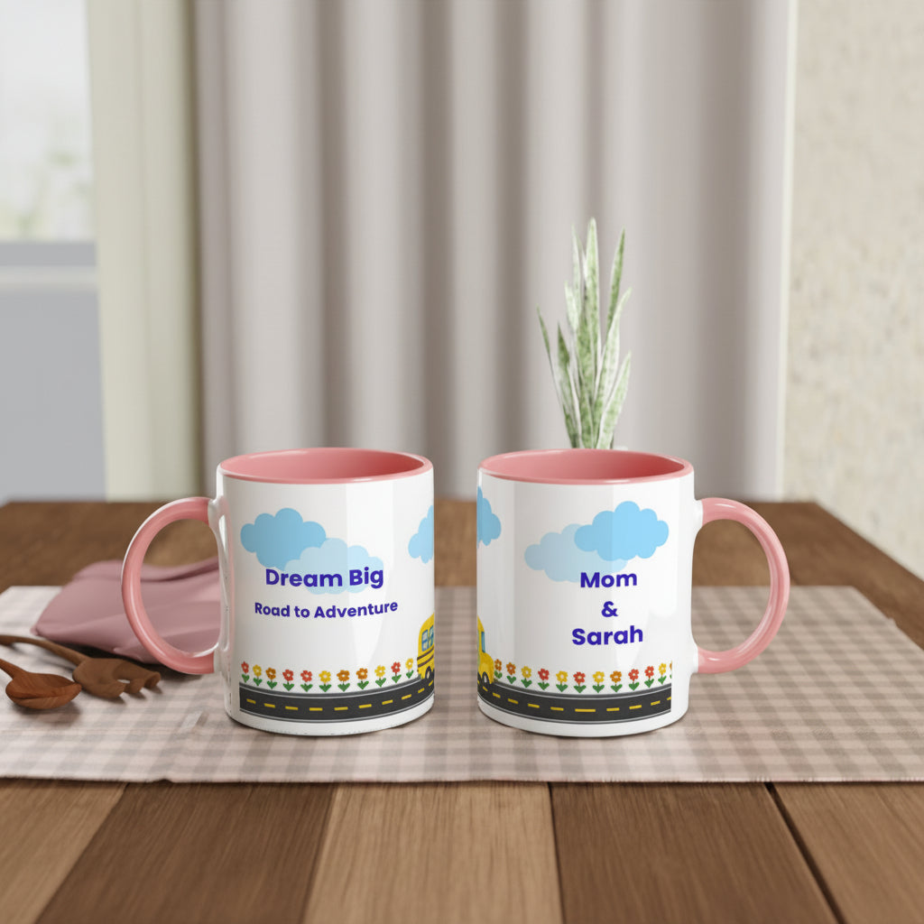 Two personalized ceramic mugs with pink interiors and handles, displayed side by side on a wooden table. The mugs feature a printed design with blue clouds, the text 'Dream Big Road to Adventure' on one mug and 'Mom & Sarah' on the other, along with a small floral road graphic.