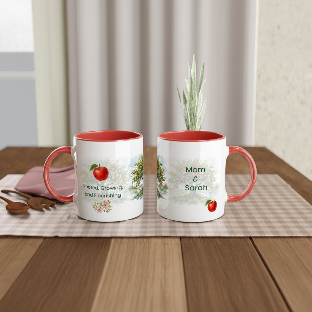 Two white ceramic mugs with coral-red handles and interiors, displayed on a wooden table. One mug shows a red apple graphic and the phrase 'Rooted, Growing and Flourishing,' while the other says 'Mom & Sarah' with a small red apple.