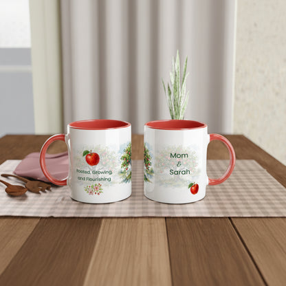 Two white ceramic mugs with coral-red handles and interiors, displayed on a wooden table. One mug shows a red apple graphic and the phrase 'Rooted, Growing and Flourishing,' while the other says 'Mom & Sarah' with a small red apple.