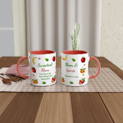 Two personalized ceramic mugs with coral red interiors and handles, displayed side by side on a checkered placemat over a wooden table. The left mug is printed with 'Sweetest Mom' and fruit illustrations, while the right mug is printed with 'Mom & Sarah' and 'Rooted in Love' with more fruit illustrations.
