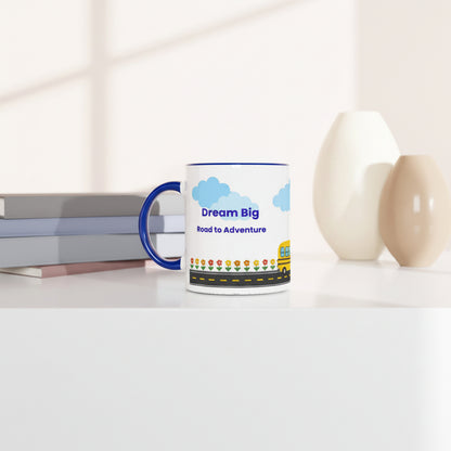 A white ceramic mug with a blue handle and blue rim, featuring a colorful graphic that reads 'Dream Big Road to Adventure' with clouds, flowers, and a yellow school bus. The mug is styled on a white surface next to books and two neutral-colored vases.