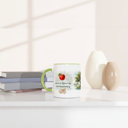 A white ceramic mug with a green handle and green rim, featuring a red apple illustration and the words 'Best Growing and Flourishing' on a white surface next to stacked books and two ceramic vases.