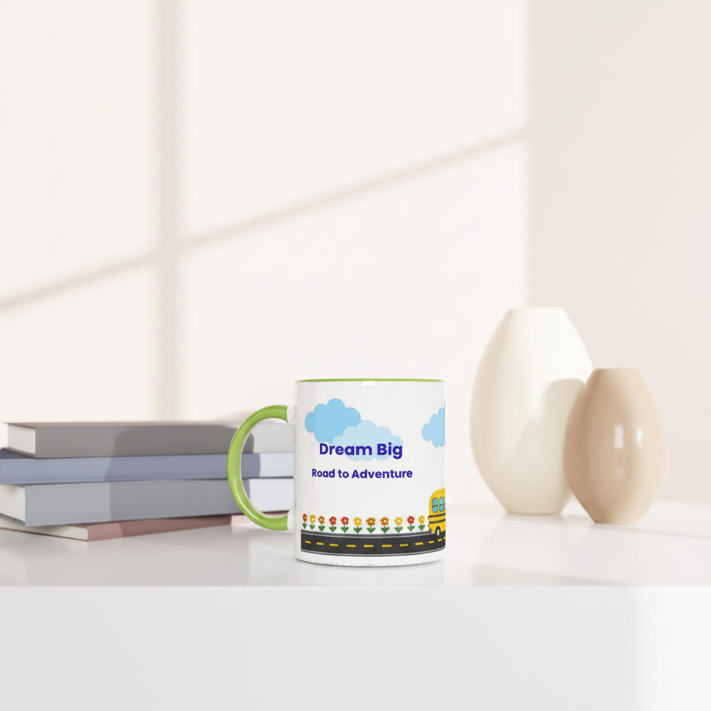 A white ceramic mug with a green handle sits on a white table in a bright, modern room. The mug features a colorful graphic of blue clouds, a yellow school bus, and the text 'Dream Big Road to Adventure'.