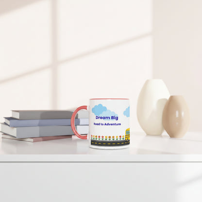 A white ceramic mug with a red handle and rim sits on a white table next to stacked books and two ceramic vases. The mug features a colorful illustration of a road, clouds, and the text 'Dream Big Read to Adventure'.