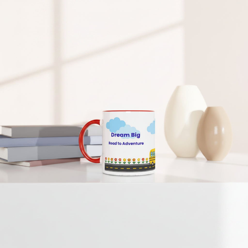 A white ceramic coffee mug with a red handle and rim sits on a white table. The mug features a colorful graphic with blue clouds, the text 'Dream Big Road to Adventure', and a yellow school bus.