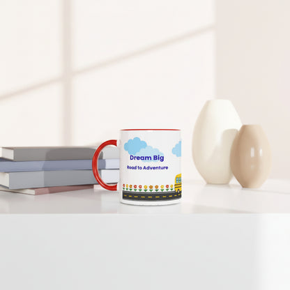 A white ceramic coffee mug with a red handle and rim sits on a white table. The mug features a colorful graphic with blue clouds, the text 'Dream Big Road to Adventure', and a yellow school bus.