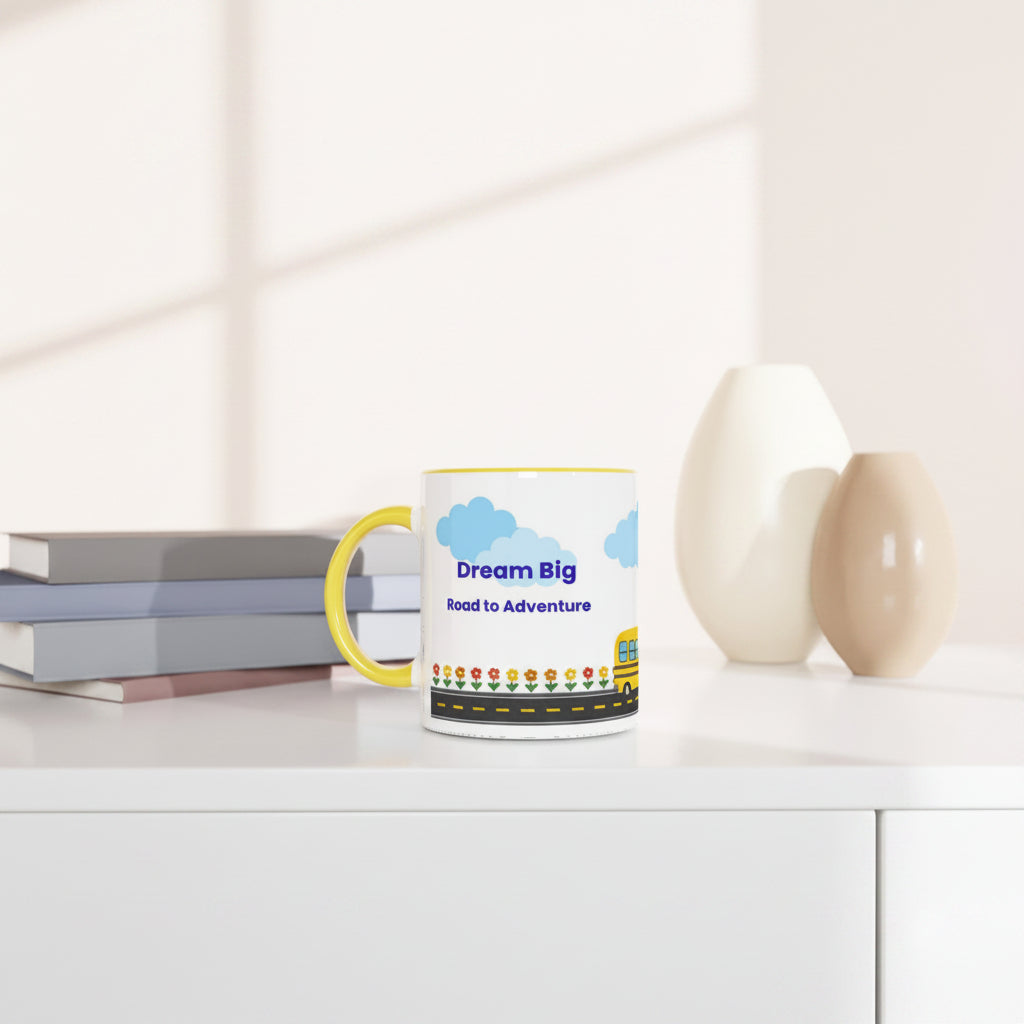 A white ceramic mug with a yellow handle sits on a white table in a bright, modern room. The mug features a colorful graphic with blue clouds, the text 'Dream Big Road to Adventure', and a yellow school bus.