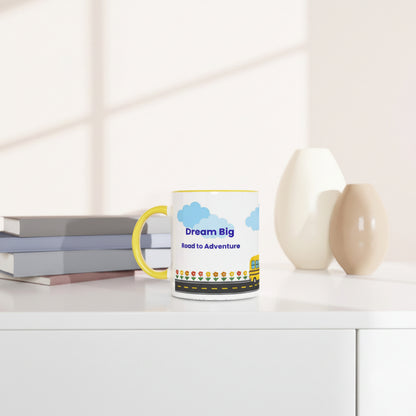 A white ceramic mug with a yellow handle sits on a white table in a bright, modern room. The mug features a colorful graphic with blue clouds, the text 'Dream Big Road to Adventure', and a yellow school bus.