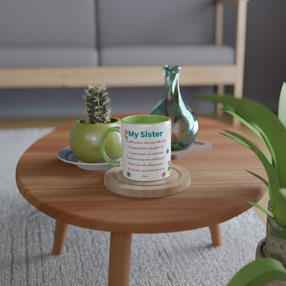 A personalized ceramic mug with a green handle and interior sits on a wooden coaster on a round wooden side table. The mug has a white exterior with colorful text and small floral accents.