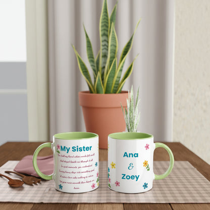 Two white ceramic mugs with light green interiors and handles are displayed on a wooden table in a styled indoor setting. One mug shows the text 'My Sister' with a poem, and the other shows 'Ana & Zoey' with small colorful flowers.