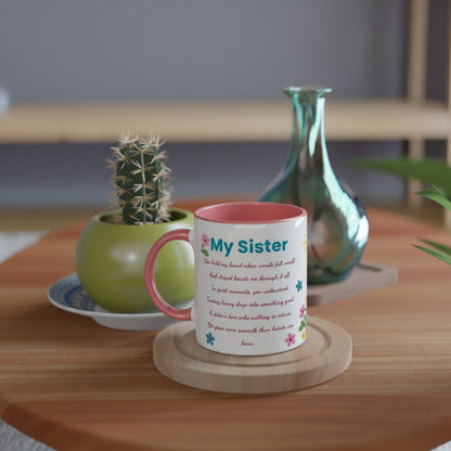 A white ceramic mug with a pink interior and handle, printed with a poem titled 'My Sister' and small colorful flowers, sitting on a wooden coaster on a wooden table. A small potted cactus and a green glass vase are in the background.