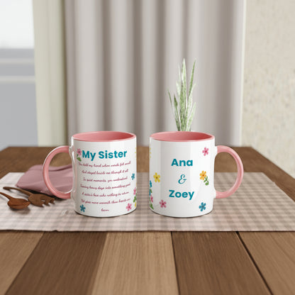 Two personalized ceramic mugs with pink interiors and handles, displayed on a wooden table with a checkered runner, a small plant, and a pink plate in the background. One mug reads 'My Sister' with printed text, and the other reads 'Ana & Zoey' with small colorful flowers.