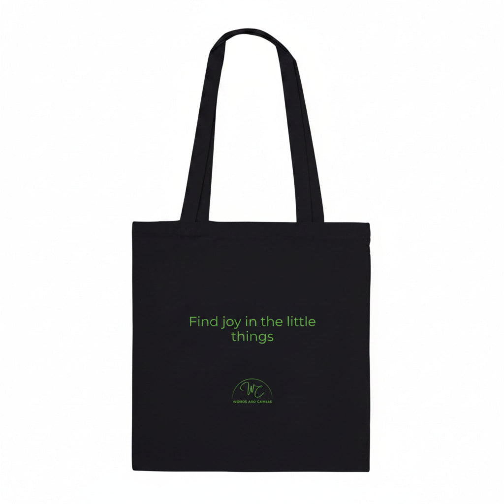 A black canvas tote bag with two shoulder straps, featuring the green printed text 'Find joy in the little things' and a small green circular logo near the bottom, shown against a plain white background.