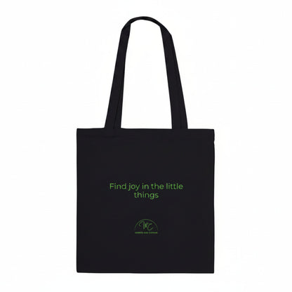 A black canvas tote bag with two shoulder straps, featuring the green printed text 'Find joy in the little things' and a small green circular logo near the bottom, shown against a plain white background.