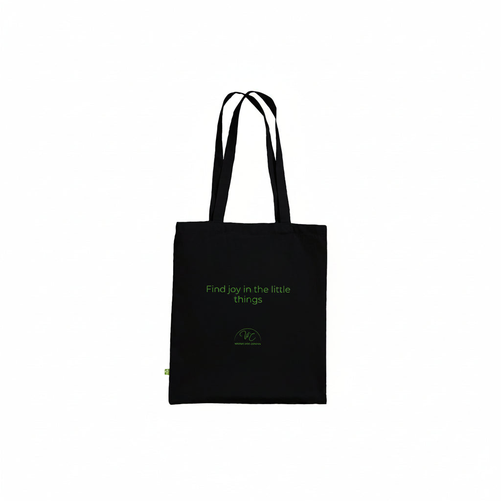 A black canvas tote bag with black shoulder straps, featuring the phrase 'Find joy in the little things' and a small green circular logo printed on the front, set against a plain white background.