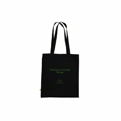 A black canvas tote bag with black shoulder straps, featuring the phrase 'Find joy in the little things' and a small green circular logo printed on the front, set against a plain white background.