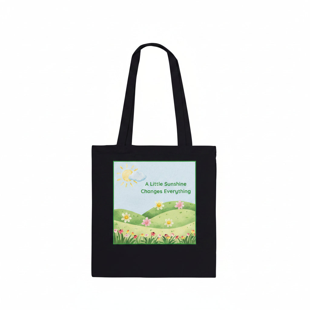 A black canvas tote bag with two shoulder straps, featuring a colorful printed graphic on the front. The graphic shows a bright yellow sun, a blue sky, green rolling hills, and a field of pink and yellow flowers with the text 'A Little Sunshine Changes Everything'.