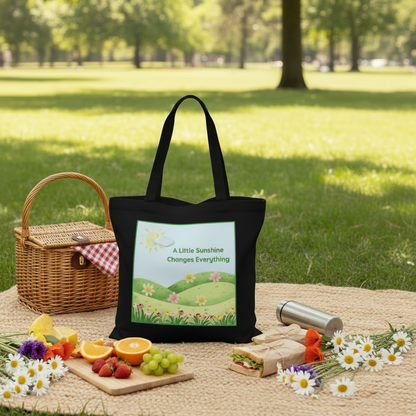 A black tote bag with a colorful landscape print reading 'A Little Sunshine Changes Everything' is set on a picnic blanket in a park. A wicker picnic basket, fruit, flowers, and a thermos are arranged around it on the grass.
