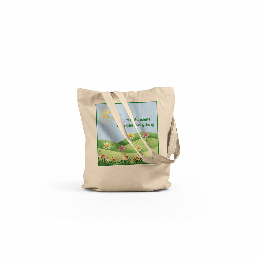 A beige canvas tote bag with a printed graphic of a sunny green field and flowers, featuring the text 'Little Sunshine changes everything' on a white background.