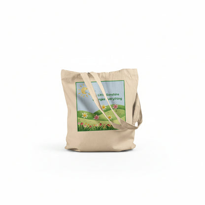 A beige canvas tote bag with a printed graphic of a sunny green field and flowers, featuring the text 'Little Sunshine changes everything' on a white background.