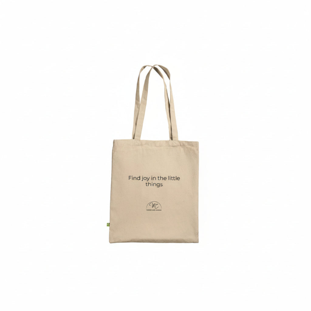 A beige canvas tote bag with black printed text reading 'Find joy in the little things' and a small circular logo near the bottom, shown on a plain white background.