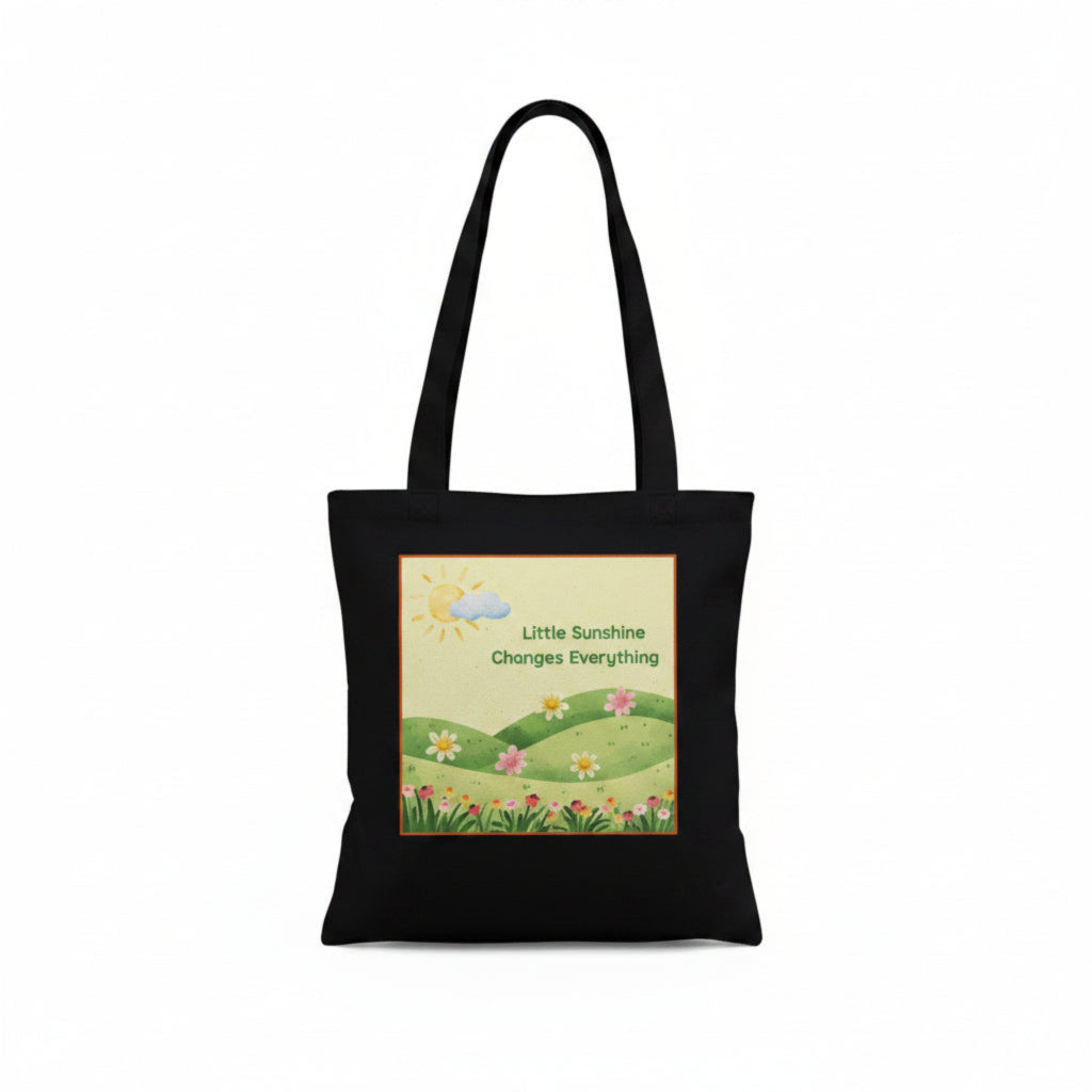 A black canvas tote bag with two shoulder straps, featuring a square graphic print on the front. The print shows a sunny landscape with green hills, small colorful flowers, a sun, and the text 'Little Sunshine Changes Everything'.