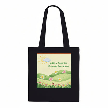A black canvas tote bag with two shoulder straps, featuring a square printed graphic on the front. The graphic shows a sunny landscape with green hills, small flowers, a yellow sun, and a blue cloud, with the text 'A Little Sunshine Changes Everything'.