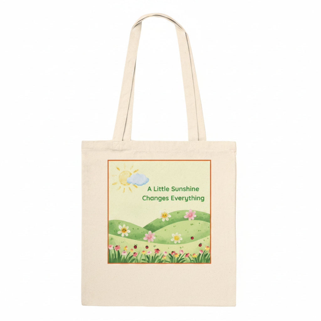 A natural canvas tote bag with a printed illustration of a sunny meadow, flowers, and the text 'A Little Sunshine Changes Everything' on the front, shown against a plain white background.