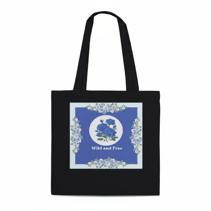 A black canvas tote bag with long handles, featuring a square graphic print on the front. The print has a blue background with a central white circle containing blue roses, surrounded by a border of light blue and white flowers, and the words 'Wild and Free' printed below.