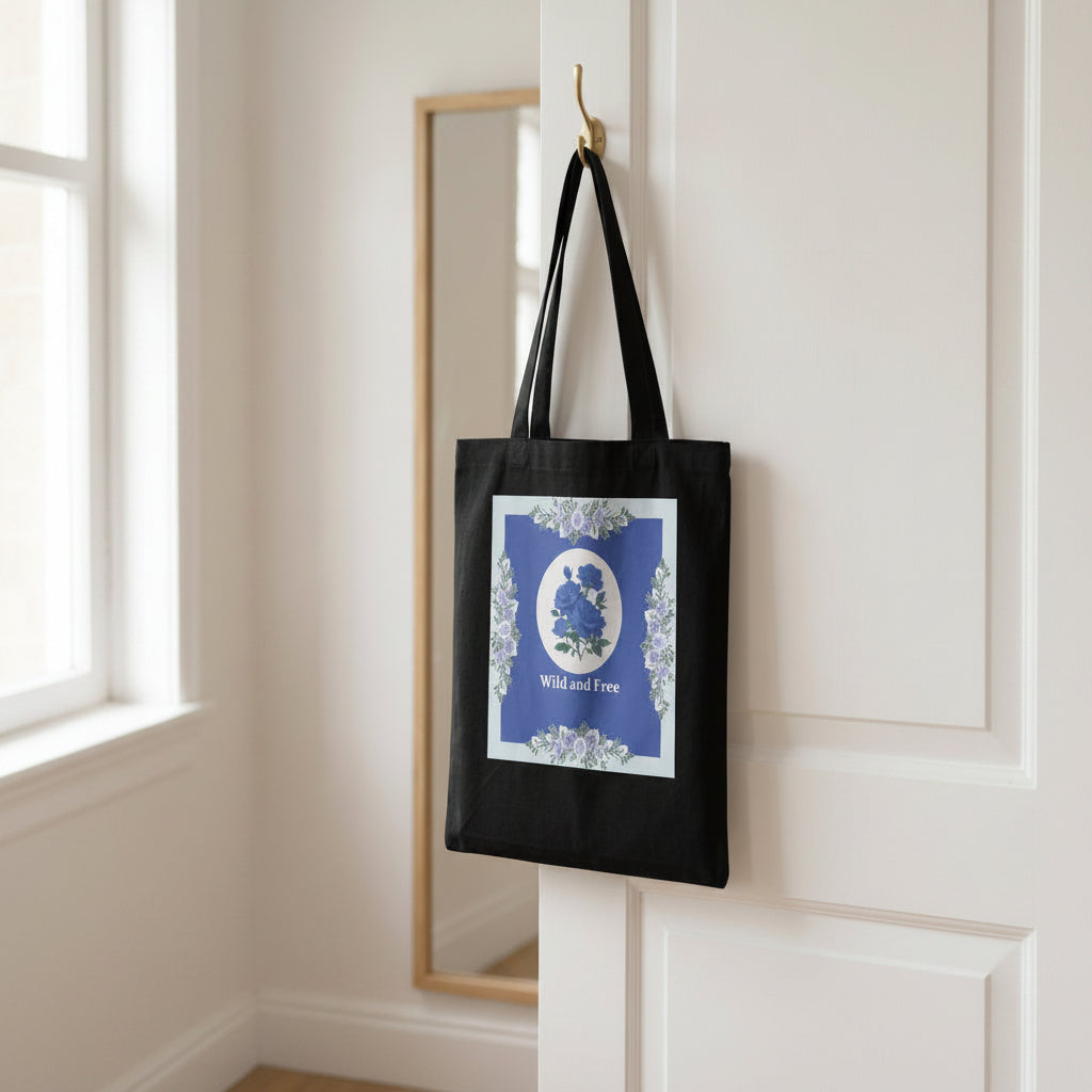 A black canvas tote bag hanging from a gold hook on a white door. The bag features a large blue and white graphic print with blue wildflowers, a white oval, and the text 'Wild and Free' surrounded by a floral border.