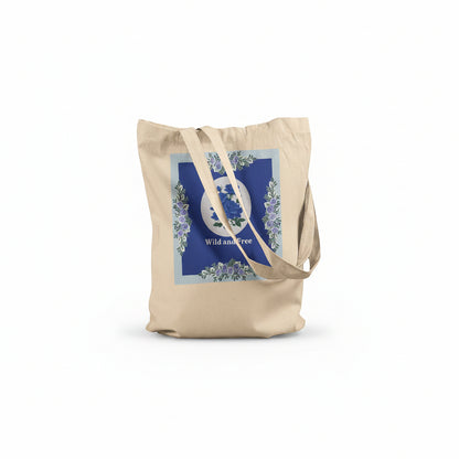 A natural canvas tote bag with a blue and white floral graphic on the front, featuring the words 'Wild and Free' and long shoulder straps, set against a plain white background.